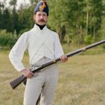 Man dressed in historical uniform with musket in a field setting.