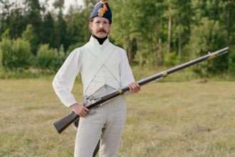 Man dressed in historical uniform with musket in a field setting.