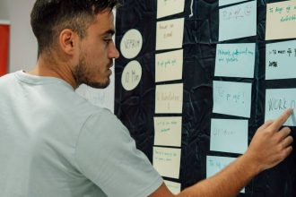 A man in a casual t-shirt organizes notes on a wall, focusing on a 'WORK OUT' sign.
